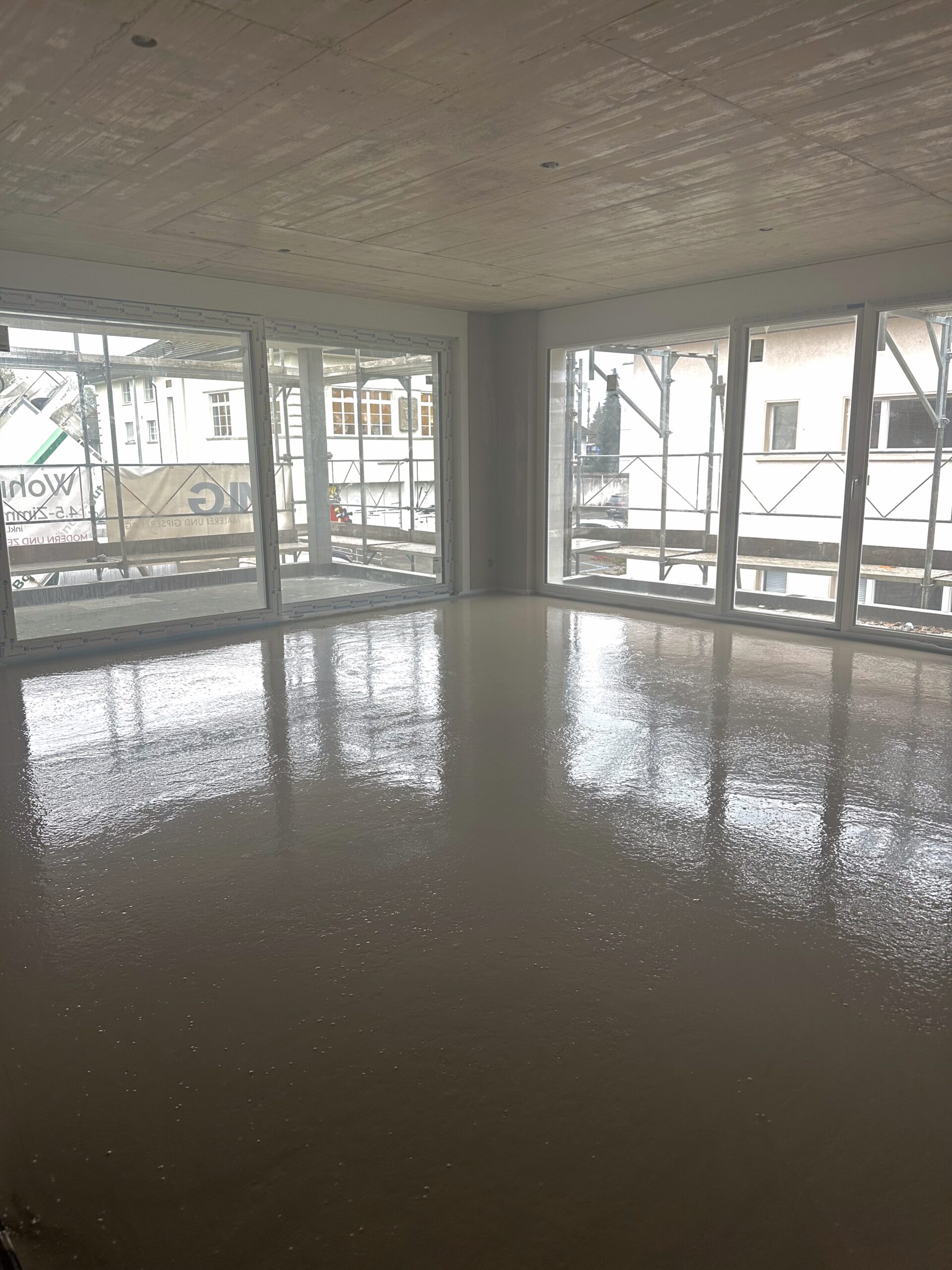 Ein Raum mit grossen Fensterfronten, durch die ein Gerüst und benachbarte Gebäude sichtbar sind. Der Boden ist frisch gegossen und glänzend, die Decke ist aus Beton mit sichtbaren Schalungsspuren.